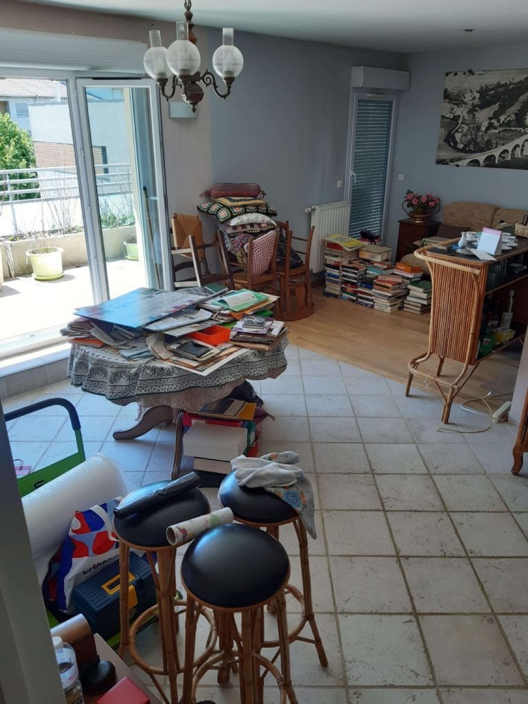 Pièce de vie encombrée de piles de livres, meubles et tabourets avant débarras complet.