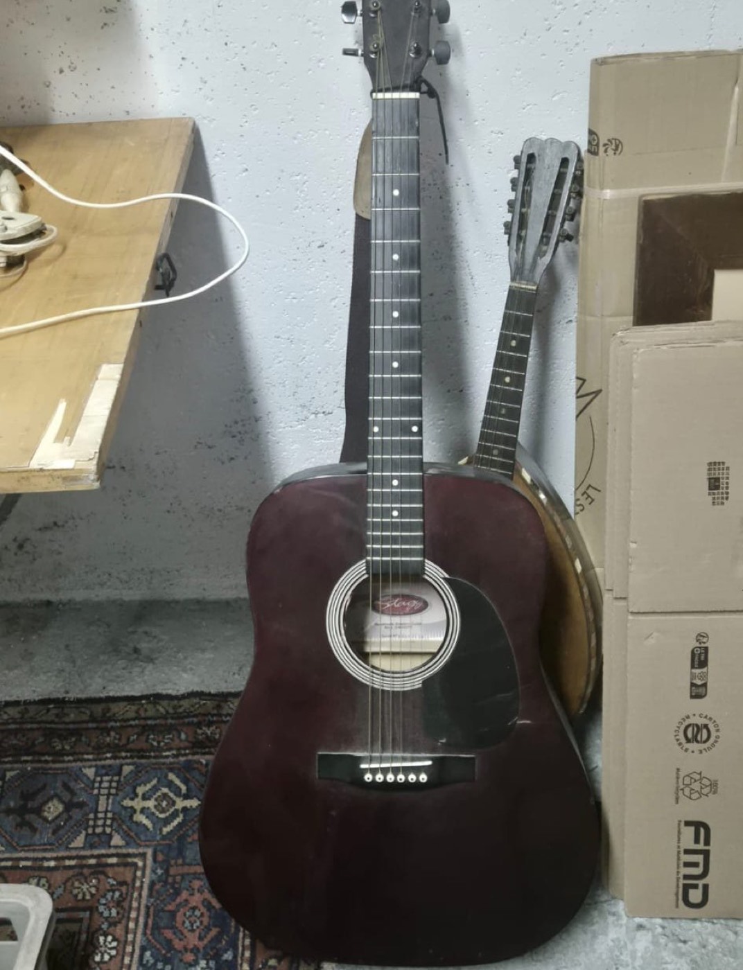 Guitare folk acoustique Stagg de couleur bordeaux posée contre un mur, avec une mandoline en arrière-plan